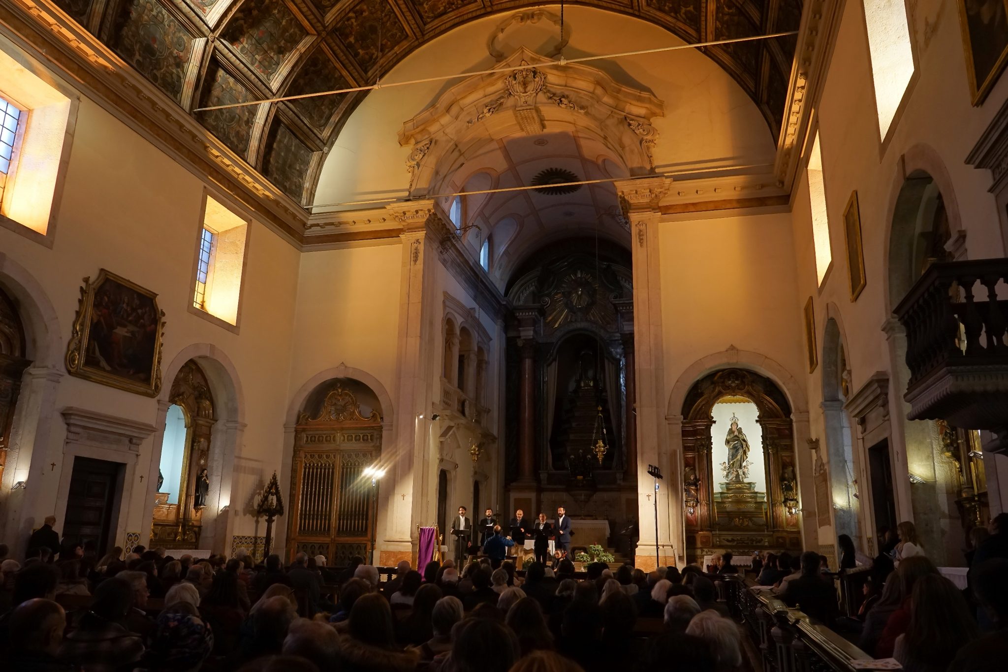Interior of a church with a choir acting next to the altar and several people seated in front of it. These are with their backs to the observer