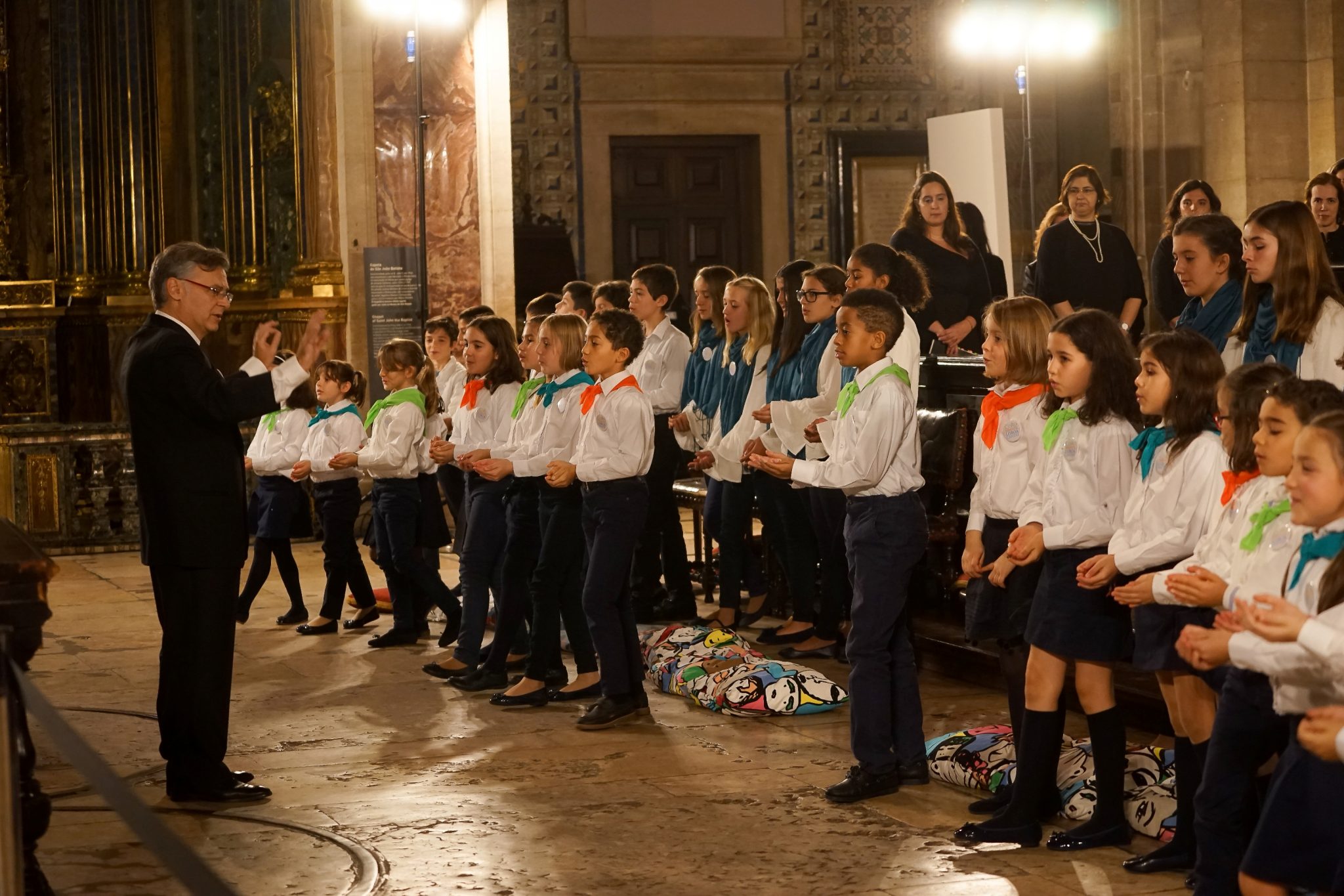 Several children, sideways to the viewer, wearing white shirts and colorful scarves around their necks, sing in front of the conductor (left of image)