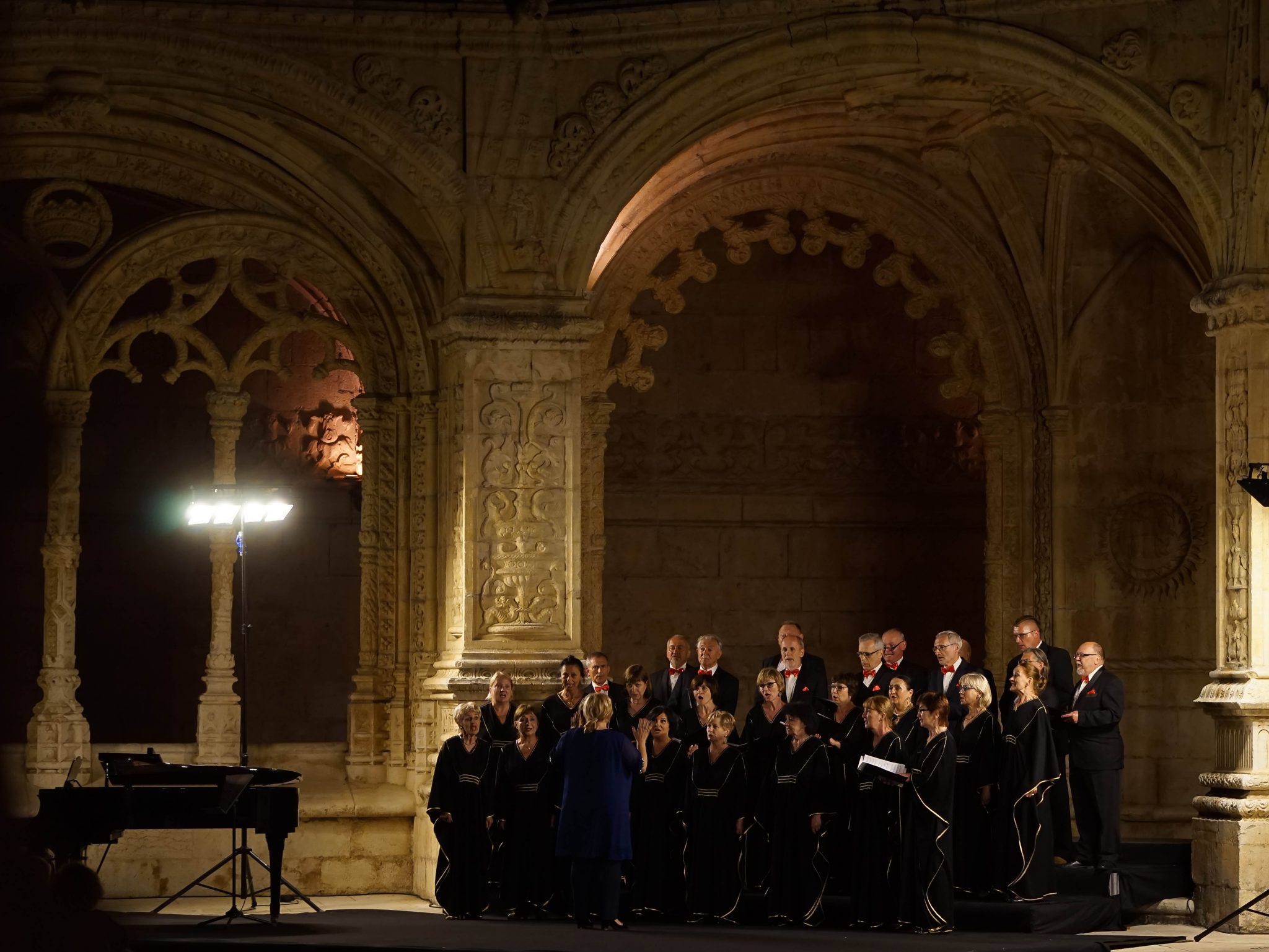 Coro composto por homens e mulheres, vestidos com roupas escuras no Claustro do Mosteiro dos Jerónimos à noite