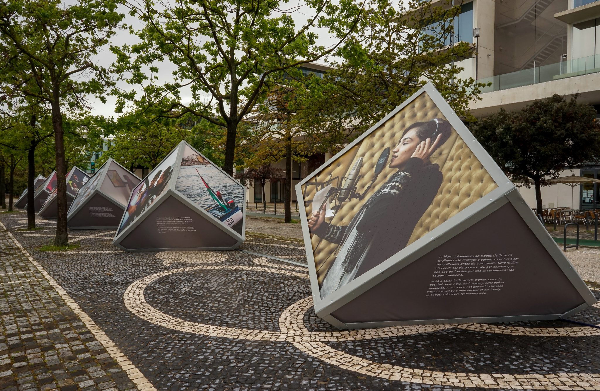 Perspectiva diagonal de uma sequência de grandes cubos pousados na calçada portuguesa de uma alameda do Parque das Nações, exibindo nas suas diferentes faces fotografias de mulheres palestinianas de Gaza que integram a exposição de Monique Jaques.