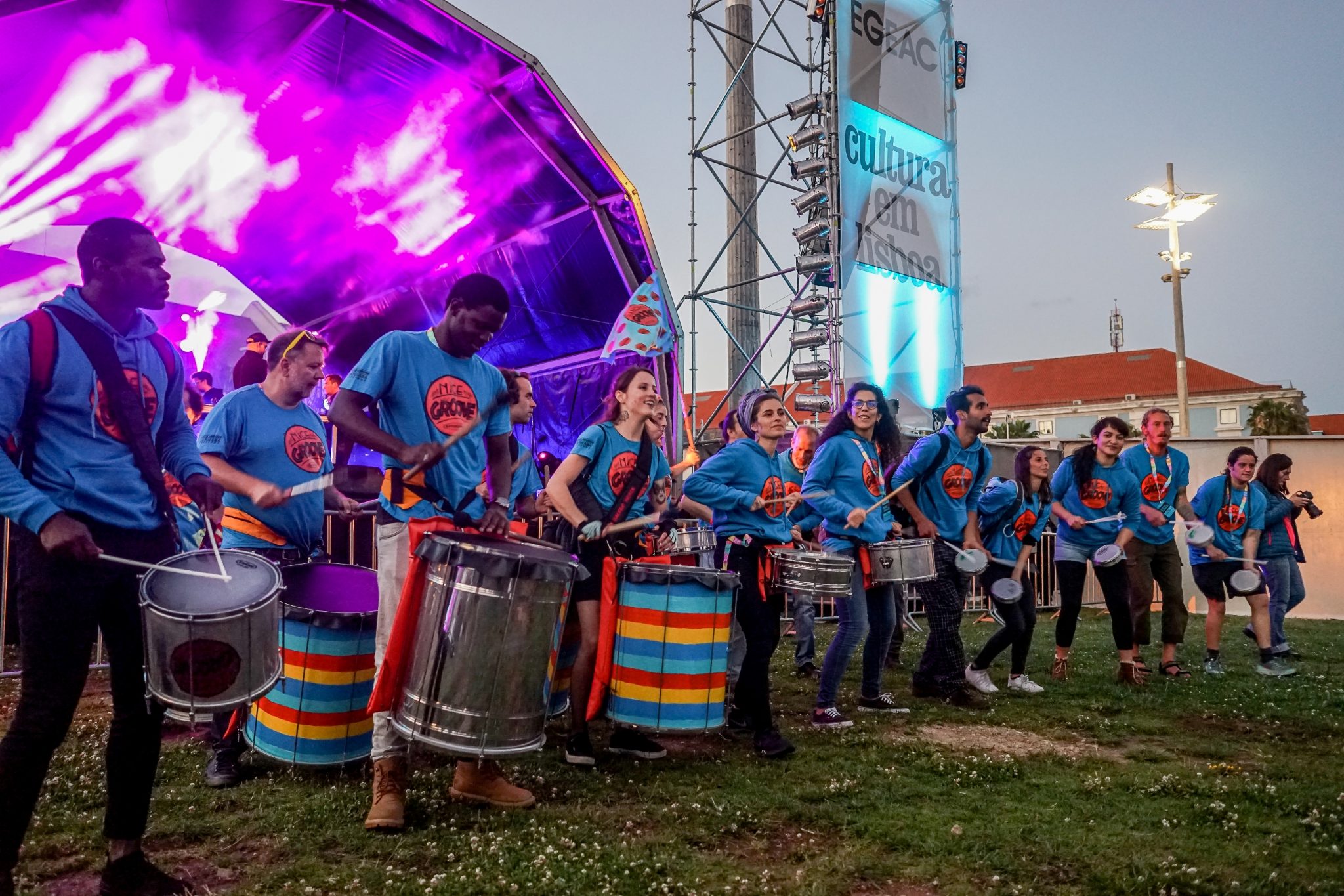 Grupo de homens e mulheres com tambores. Vestem camisolas azuis e atuam em frente a um palco com luzes roxas. Luz de fim de dia