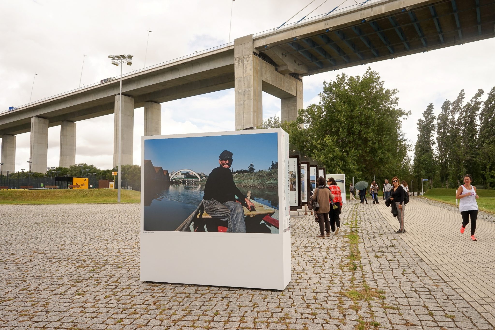 Em primeiro plano, uma estrutura com uma fotografia de um pescador no Rio Trancão. Em segundo plano, várias pessoas a olharem para outras fotografias e a Ponte Vasco da Gama.