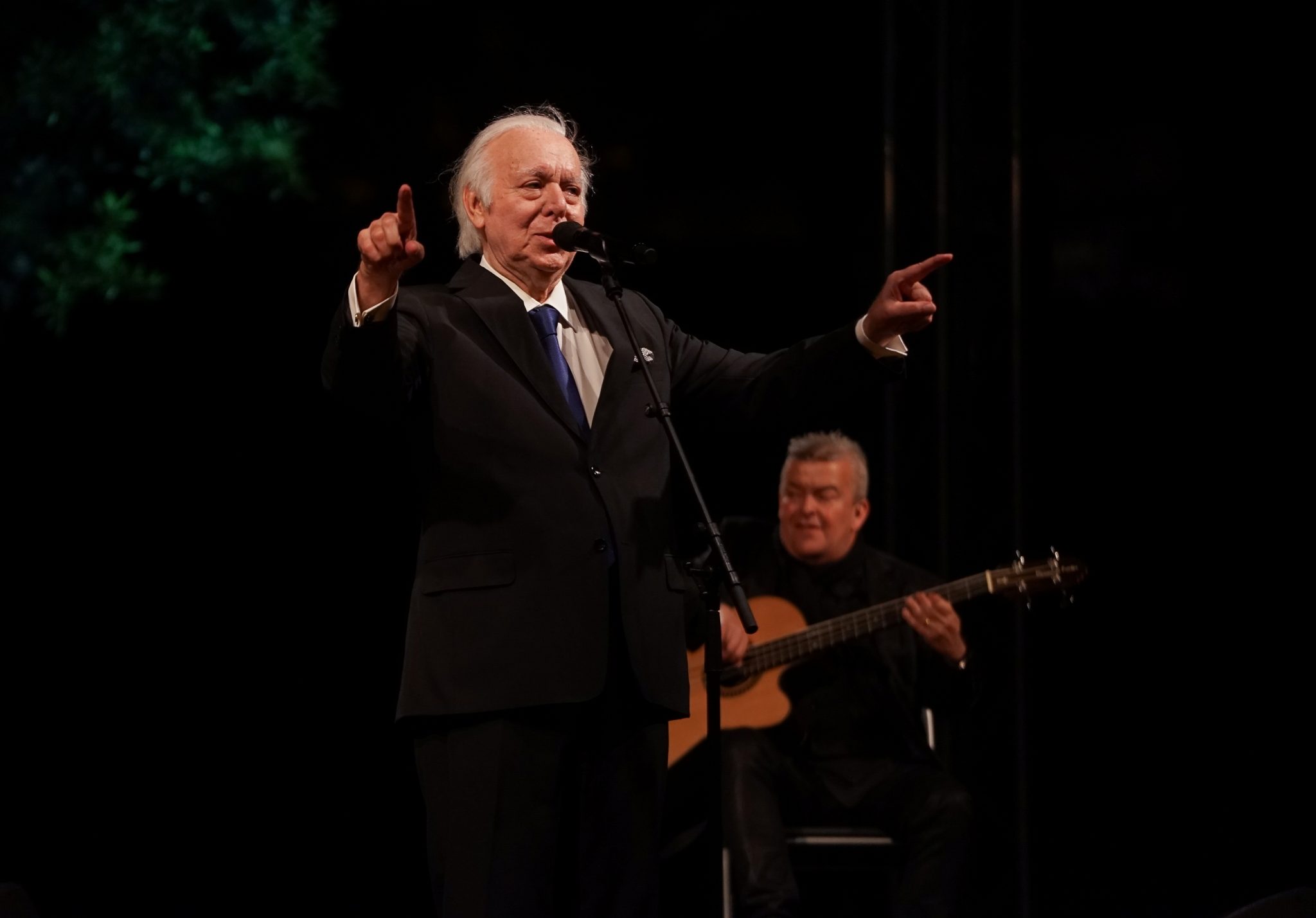 Fadista Carlos do Carmo em palco, diante do microfone, a cantar de braços abertos e dedos indicadores esticados, de frente para o observador