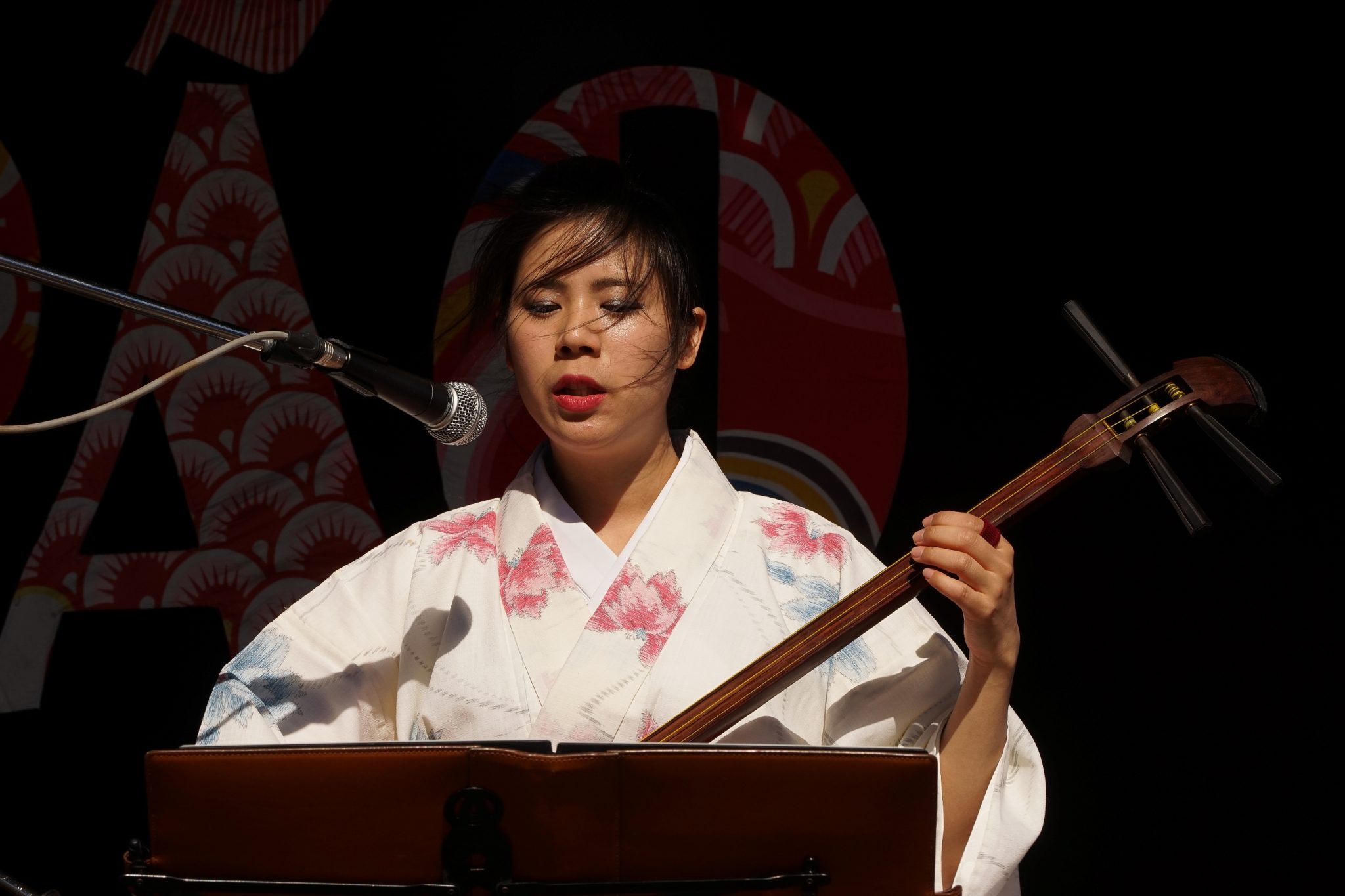 De frente para o observador, mulher com trajes japoneses toca Shamisen em frente a um microfone