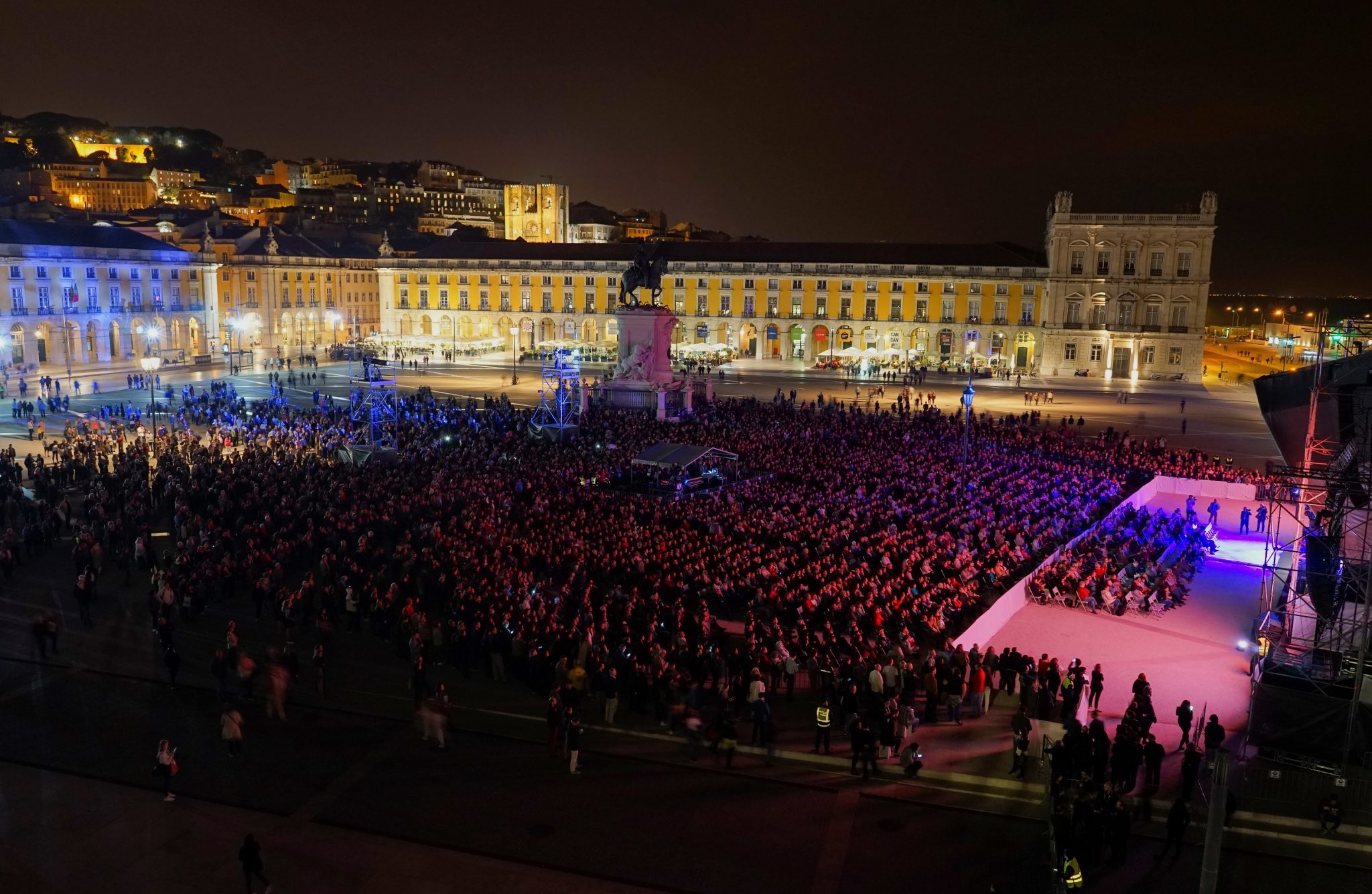 Vista noturna de uma multidão de pessoas na Praça do Comércio iluminada, encimada, do lado esquerdo, pela colina do Castelo de S. Jorge.