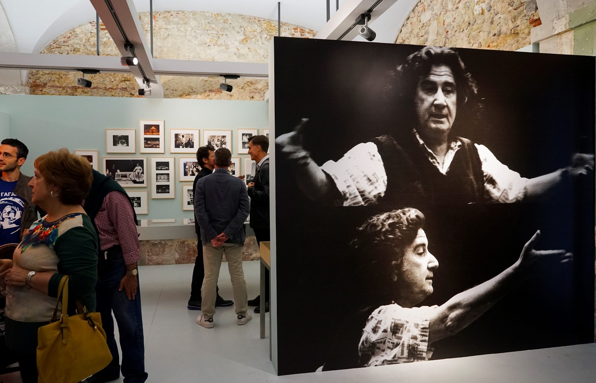Algumas pessoas, em pé, pontuam uma exposição de fotografias com uma imagem de grande formato, a preto e branco, em destaque no lado direito.