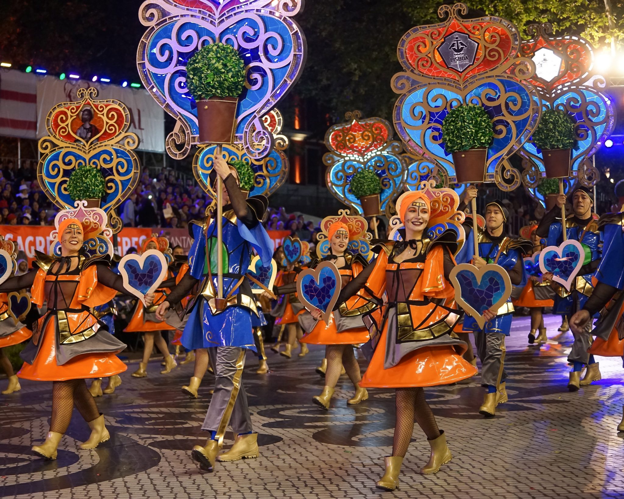 Um grupo de marchantes composto por homens e mulheres, elas com roupas cor-de-laranja e eles de roupas azuis e arcos altos nos braços, avançam para a esquerda da imagem enquanto sorriem.