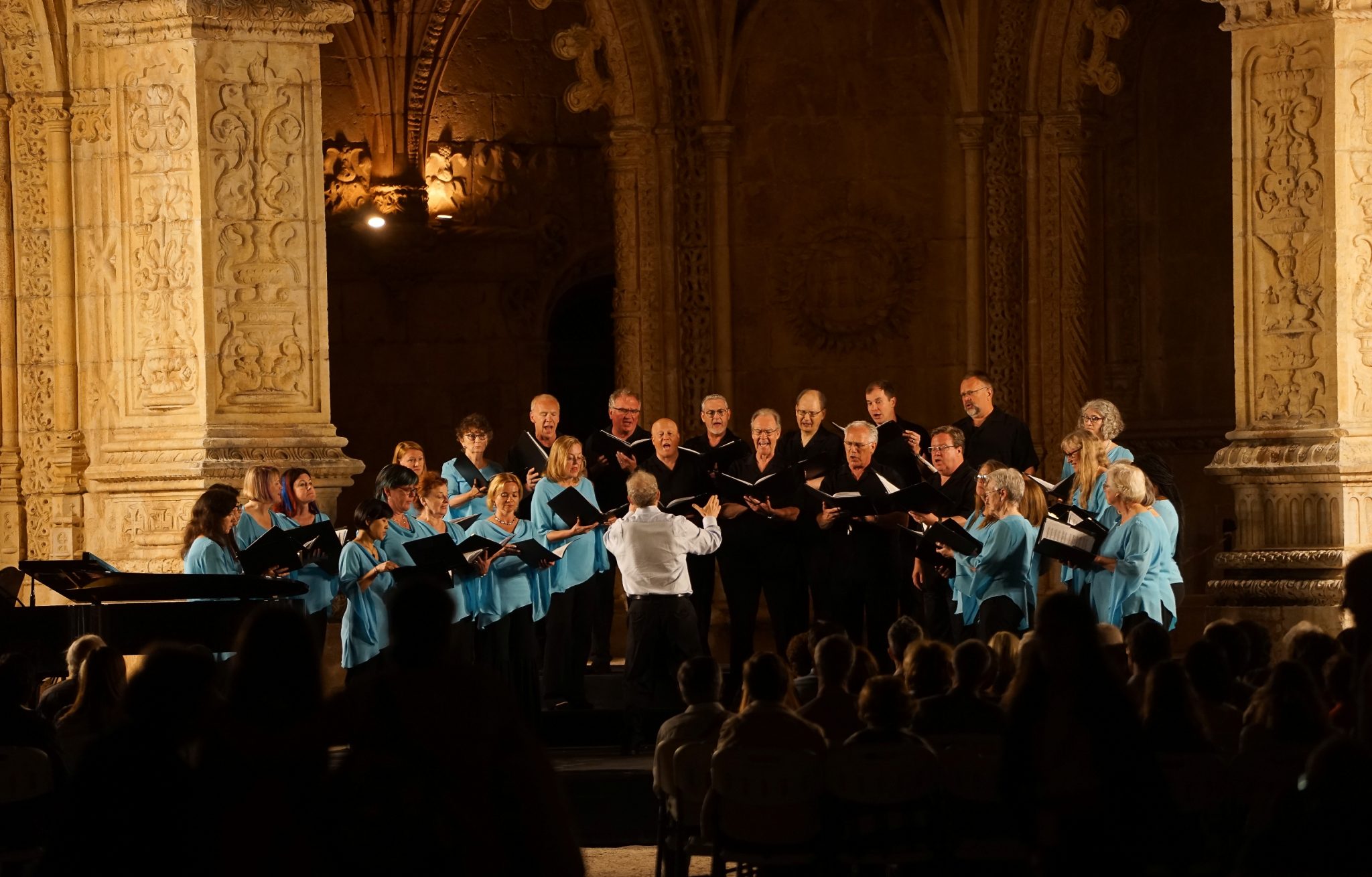 Cerca de vinte cantores do coro Soli Deo Gloria (homens vestidos de negro, as mulheres com blusa de azul claro) cantam no Claustro do Mosteiro dos Jerónimos. Ao centro encontra-se o maestro, de camisa branca.